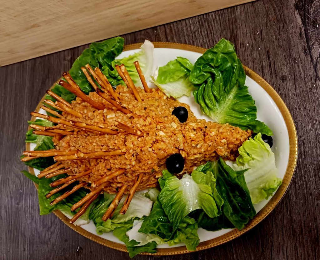 Serviervorschlag eines veganen Mettigels. Angerichtet mit Salatblättern auf einer Platte mit Goldrand.